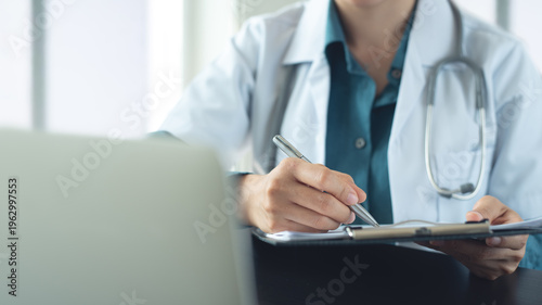 Doctor writing on medical document, medical report on clipboard with laptop on office desk