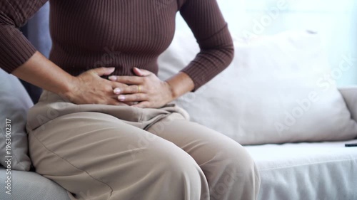 Close-up of a woman sitting on a sofa clutching her stomach in pain, suffering from abdominal cramps, menstruation, or digestive issues at home. Concept of female health and stomachache