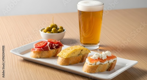 Assortment of Spanish tapas appetizers served on a white rectangular plate alongside a glass of frothy beer and olives.