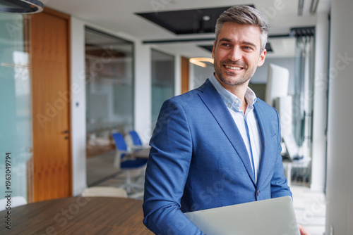 Professional businessman smiling and holding a laptop, standing in a brightly lit office corridor, representing leadership and success