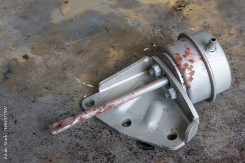 Tableau sur toile A metal turbocharger wastegate actuator with a rusty control rod and mounting bracket is shown from a top-down view on a dirty industrial floor for engine part replacement