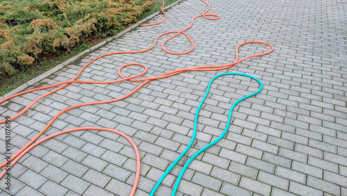 Outdoor scene featuring winding orange and teal garden hoses lying messy on a grey cobblestone driveway, representing home irrigation, gardening, and household tasks