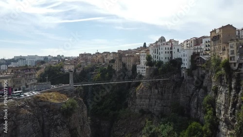 Ground-level aerial drone view of the Sidi M'Cid suspension bridge and the steep limestone cliffs of Constantine, Algeria, under a bright sky.
