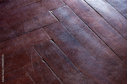 Close-up of a dark brown herringbone parquet wood floor showing signs of wear, scratches, and scattered dust particles under bright natural lighting