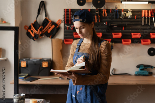 Female small business owner in overalls planning furniture production and reading notebook in workshop