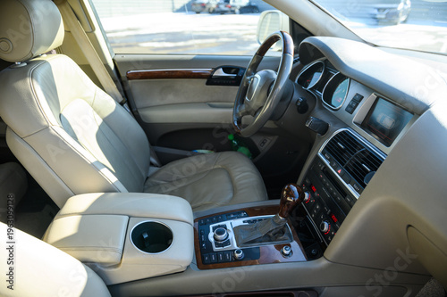 Luxury car interior with beige leather seats and wooden steering wheel. High-angle view of automotive cockpit featuring center console, gear selector, and dashboard in bright daylight.