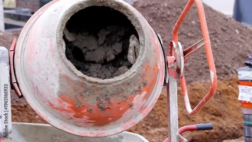 Concrete mixer drum rotates, mixing cement and aggregate, with visible construction site background and piles of earth in the vicinity, showcasing the mixing process