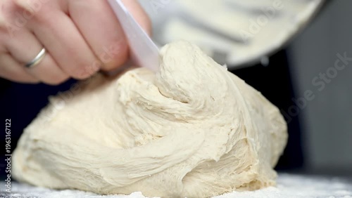 Hand of a Caucasian baker kneads soft dough with a spatula on a floured countertop, showcasing the texture and consistency of the mixture in a kitchen setting