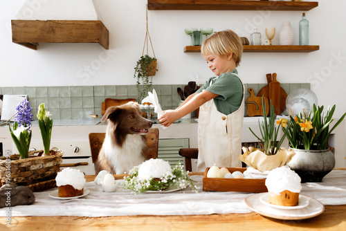 Little blonde boy with a border collie dog at a festive Easter table. Child and pet in a cozy kitchen with Easter eggs, spring flowers, and traditional cake. Happy holiday atmosphere