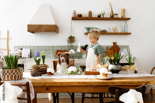 Little blonde boy with a border collie dog at a festive Easter table. Child and pet in a cozy kitchen with Easter eggs, spring flowers, and traditional cake. Happy holiday atmosphere