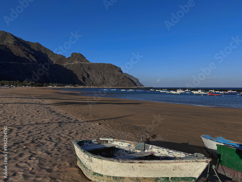 A picturesque landscape showing Las Teresitas beach in Tenerife, Canary Islands. Bright golden sand in the foreground with old traditional fishing boats, turquoise ocean with more anchored boats.