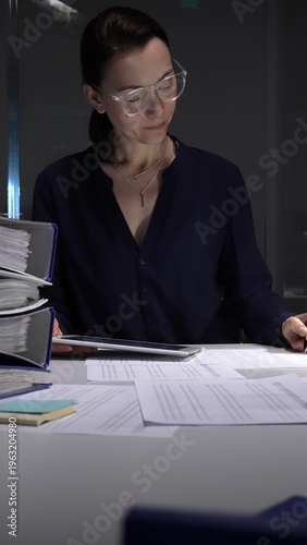 Фотография Woman working late night in office, multitasking with a tablet and writing on documents, managing heavy paperwork load