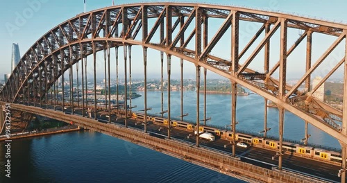 Sydney Harbour Bridge steel arch with cars and train riding across the water with city skyline and waterfront buildings in the background, modern cityscape view at daytime. Aerial drone static footage