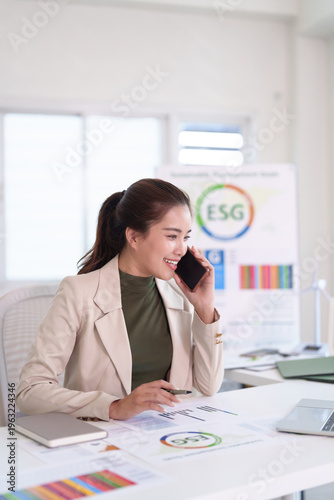 Asian businesswoman discusses ESG strategies on phone in modern office workspace analyzing sustainability data documents
