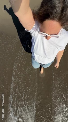 Woman standing joyfully at the beach, capturing a selfie while wearing stylish white sunglasses. Waves splash around her feet, adding a touch of summer fun and relaxation.