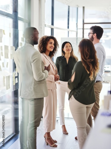 Four diverse professionals in business attire engage in discussion inside a modern office space with large windows and sticky notes on the wall