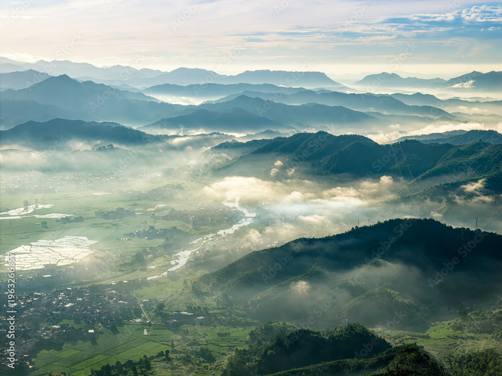 Obraz premium Panoramic aerial view of misty mountain range and rural village nestled in green valley at sunrise, highlighting serene natural beauty and peaceful morning atmosphere.