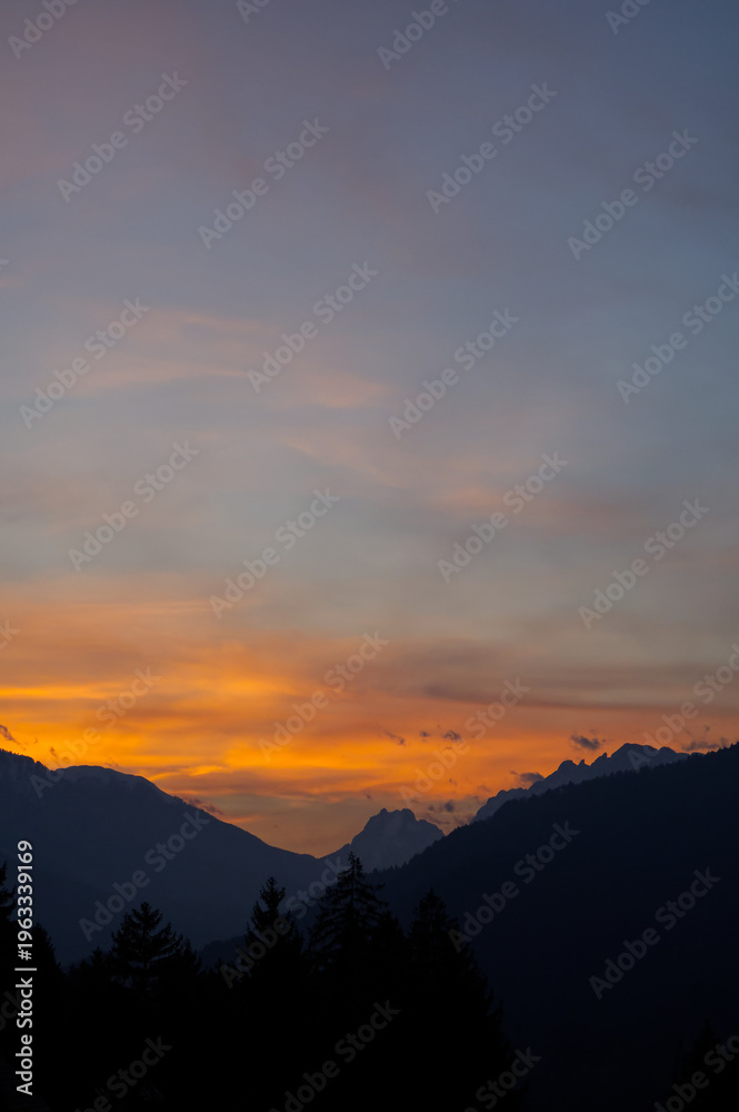 Obraz premium Spectacular sunset over a dark mountain range with silhouetted pine trees. Vibrant orange and yellow sky contrasting with dark alpine peaks. Mountain sunset background wallpaper
