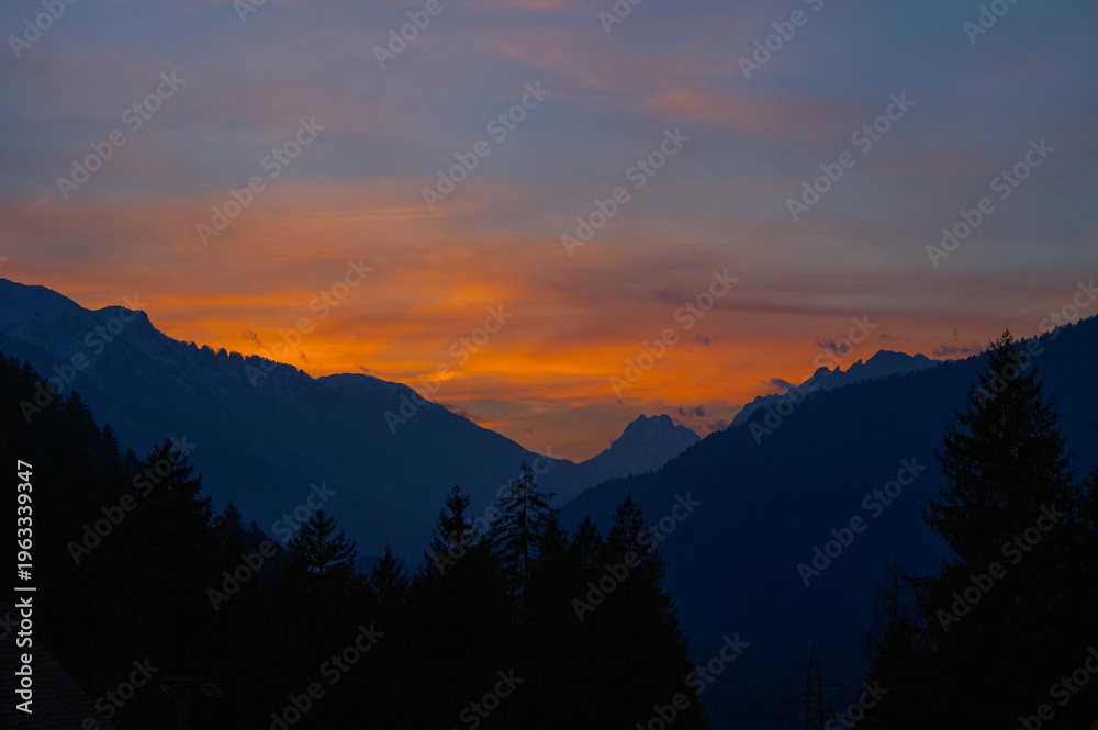 Obraz premium Spectacular sunset over a dark mountain range with silhouetted pine trees. Vibrant orange and yellow sky contrasting with dark alpine peaks. Mountain sunset background wallpaper