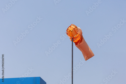 Billede på lærred Orange wind sock against blue summer sky with factory background.