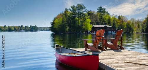 Foto Two Adirondack chairs and a red canoe on a weathered Ontario dock face a calm Muskoka lake, boathouse and pine island;  the heart of Canadian cottage country in summer