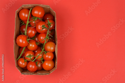 Fresh red cherry tomatoes on green vines are packed in a brown cardboard container against a solid red backdrop