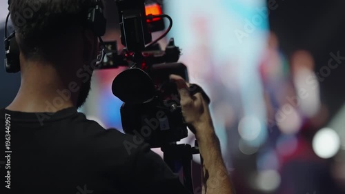 Professional videographer operating a shoulder-mounted camera with a headset during a live stage event, capturing performers under dramatic lighting with bokeh in the background.