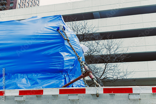 Φωτογραφία Large blue wrapped item is tied down on a truck near a parking structure in an u