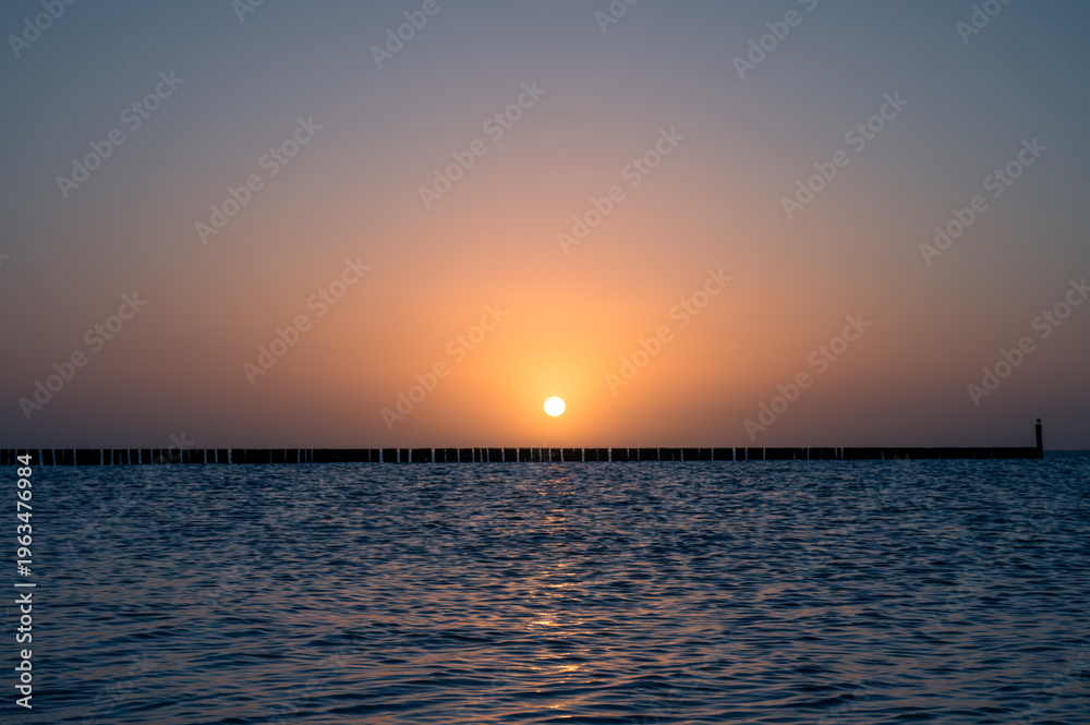 Fototapeta premium Impressive sunrise behind a row of wood over calm water, wooden groynes for coastal protection on the southern beach of 23775 Großenbrode, Baltic Sea, Germany