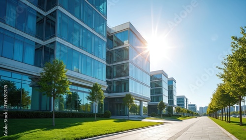 Modern glass office buildings stand on green lawn with trees. Bright sun shines on clear blue sky. This scene shows contemporary corporate architecture and landscaping.