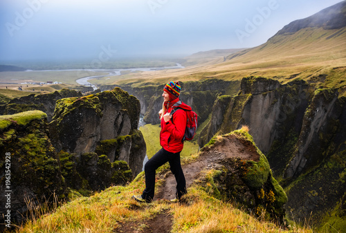 Smiling hiker with a red jacket and colorful hat stands at the edge of Iceland's Fjadrargljufur Canyon, taking in the breathtaking landscape of steep cliffs and winding river below