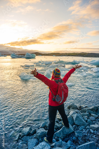 A person stands with arms raised by the ice lagoon in Iceland, celebrating the natural beauty at sunset. Icebergs float in the water, with a colorful sky overhead.