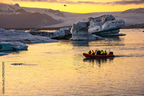 A group of tourists on a boat navigates through a glacier lagoon filled with icebergs. The golden hues of sunset reflect on the water, creating a stunning backdrop