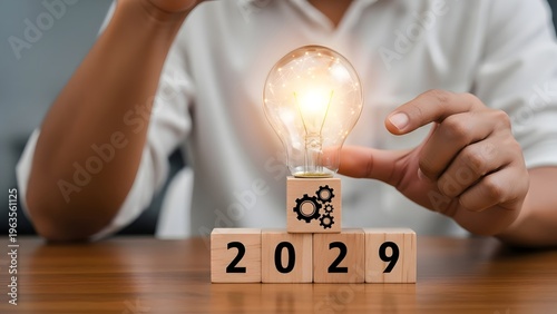 Person places a glowing light bulb on wooden blocks spelling 2029 for innovation.