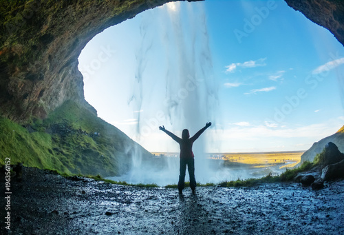 Visitor stands with arms raised in excitement behind Seljalandsfoss Waterfall in Iceland. The sun sets, illuminating the lush landscape and cascading water.