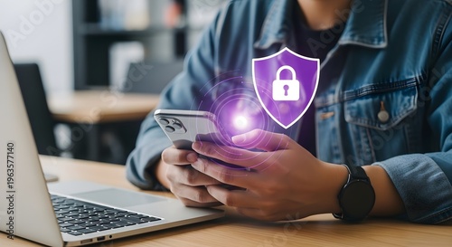 Person holding phone with purple shield and lock icon on screen near laptop