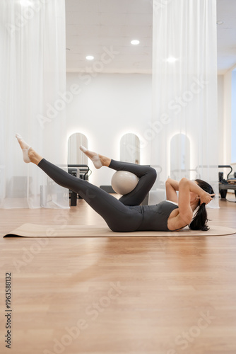 Woman in black outfit performing open-leg teaser on mat during pilates sequence in white studio. Open-leg teaser, pilates, mat, white studio, black outfit.