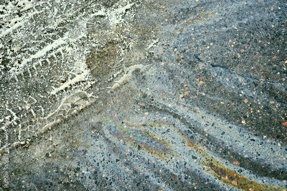 Naklejka premium Oil slick on rough asphalt road creating an iridescent rainbow pattern and showing white tire marks, indicating pollution