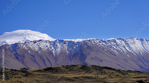The snow-capped peaks of Mount Ararat dominate the horizon under a clear blue sky, viewed across the rugged, arid landscape of Eastern Turkey