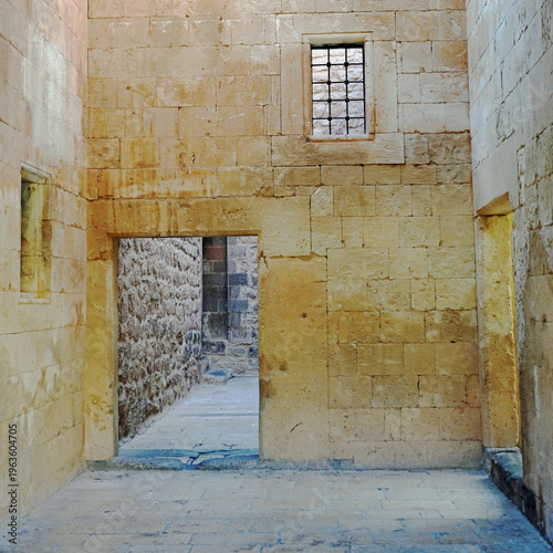 An interior view of the Ishak Pasha Palace showcases the ancient stone masonry of a sunlit courtyard, featuring a simple rectangular doorway and a high, barred window