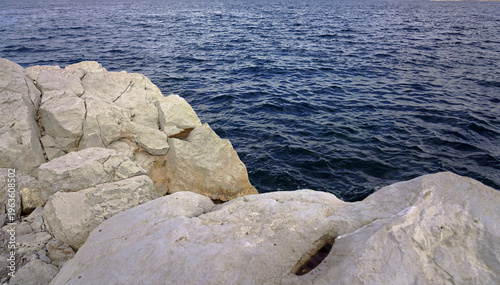 White limestone cliffs and rugged rock formations line the shoreline of Akdamar Island, meeting the clear, deep blue waters of Lake Van under a bright sky in eastern Turkey