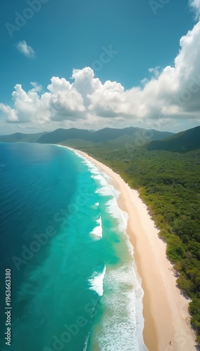 Aerial view of coastline with bright turquoise ocean waves washing onto sandy beach. Lush green forest covers hills behind the shore. Clouds drift across the clear blue sky.