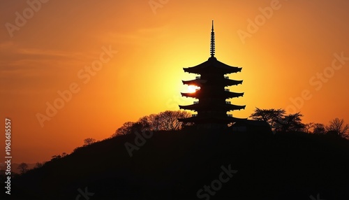 Multi-tiered pagoda silhouetted against a fiery orange sunset sky. Traditional Japanese architecture stands atop a hill with bare trees below. Golden light illuminates the scene at dusk.