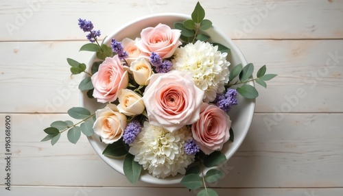 Pastel roses and white hydrangeas arranged in a white bowl on a light wood table. Ethereal floral display with eucalyptus leaves and purple accents.