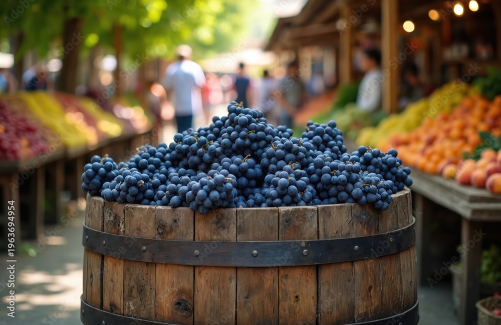 Fototapeta premium Wooden barrel brimful with dark grapes sits at outdoor market stall. Colorful fresh fruits piled high on tables behind. People mill about in background purchasing produce.