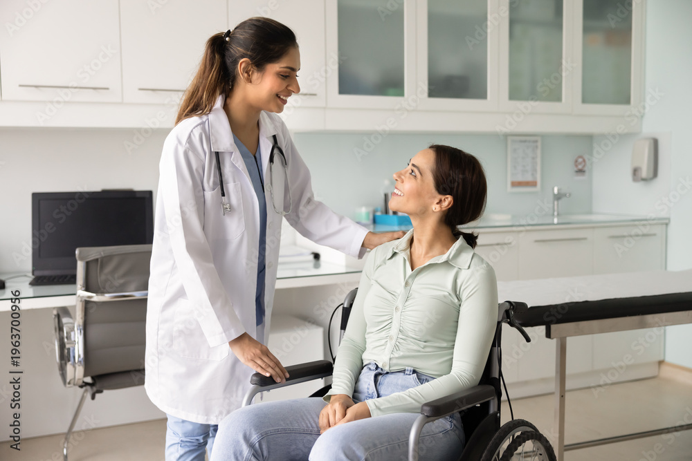 Obraz premium Indian female doctor in white coat stands beside woman patient in wheelchair, engaging in friendly conversation and offering reassurance, representing disability care, rehabilitation, medical support