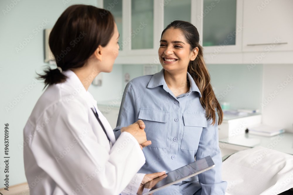 Obraz premium Professional female GP doctor in white uniform holding digital tablet shaking hands with smiling Indian woman patient at hospital office during consultation. successful treatment, healthcare support