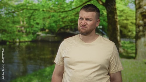 Man grimacing with visible face and stubble, wearing beige tshirt, standing in forest by a pond and small bridge among trees and grass; disgust.