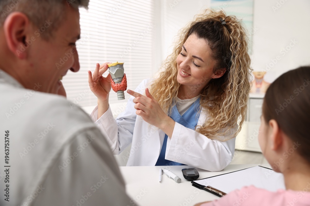 Obraz premium Smiling endocrinologist consulting man with his daughter and showing models of thyroid glands at white desk in clinic
