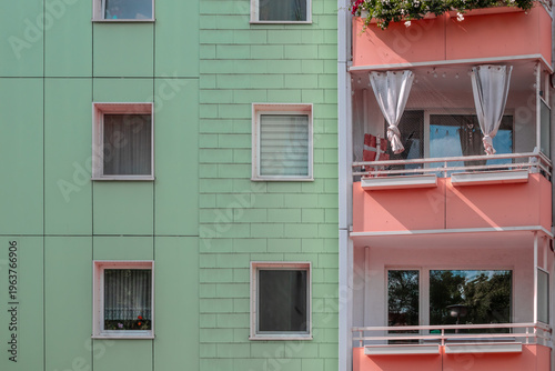 Colorful renovated East German apartment facade with mint green cladding and peach-toned balcony section, featuring windows, curtains and flower boxes, showcasing modern upgrades of classic socialist-